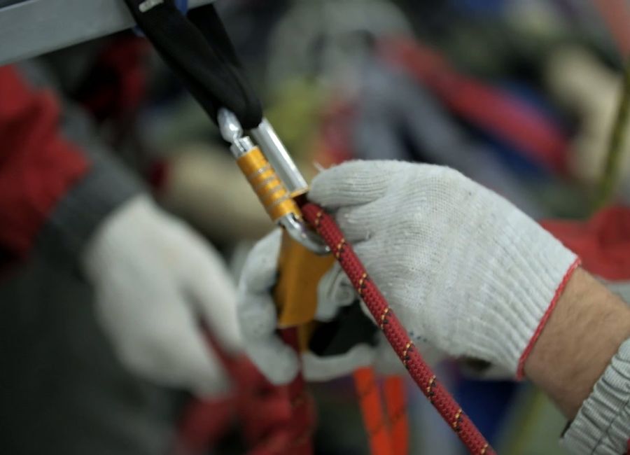Manos con guantes blancos sujetan una cuerda roja conectada a un mosquetón dorado y plateado durante un trabajo de seguridad en altura.
