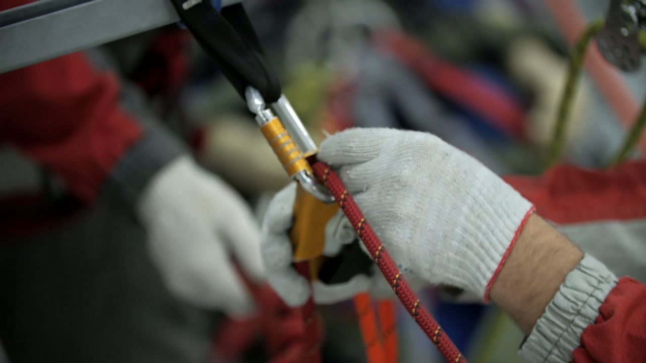 Manos con guantes blancos sujetan una cuerda roja conectada a un mosquetón dorado y plateado durante un trabajo de seguridad en altura.